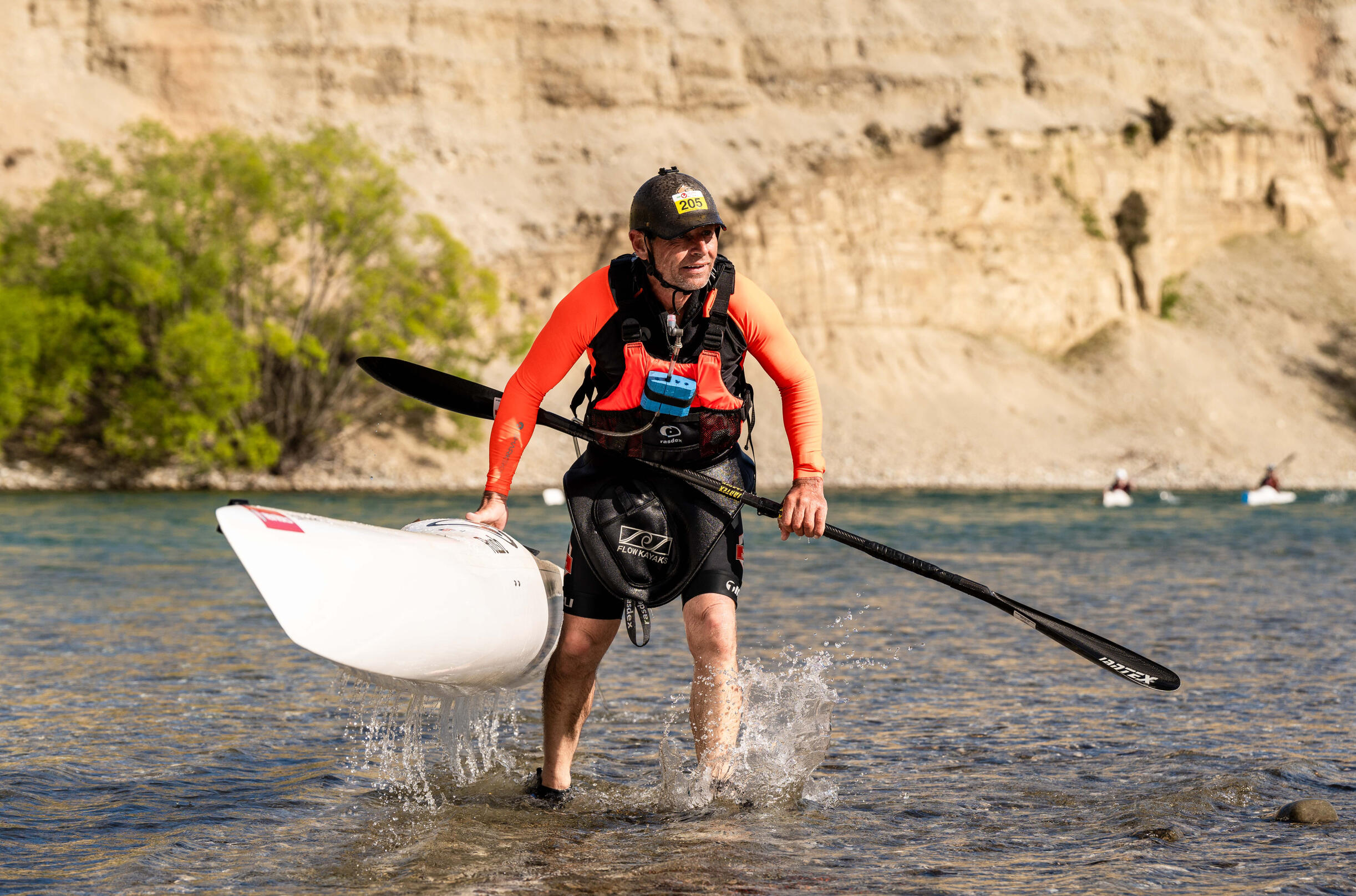 Kayaker in orange gear carrying white Kayak through shallow turquoise water, canyon cliffs behind