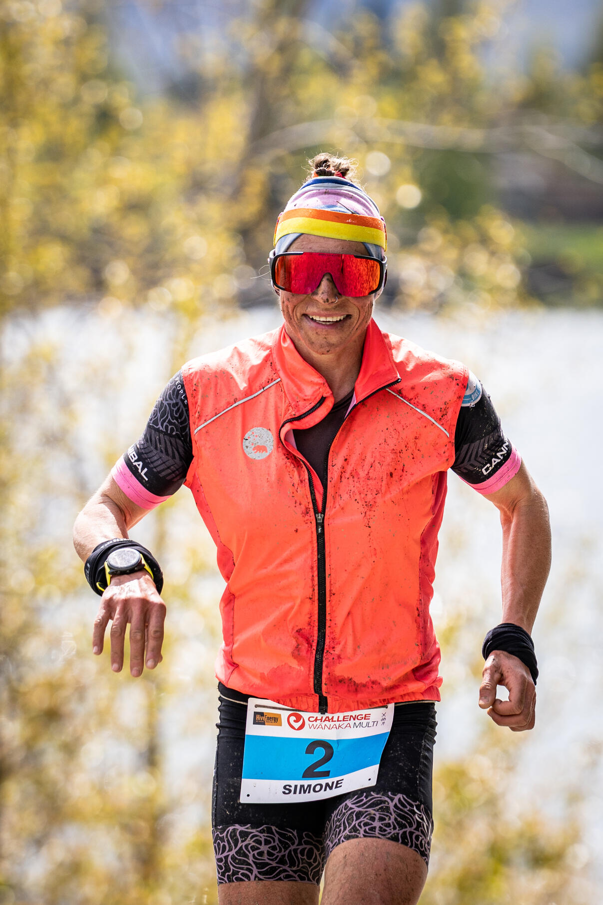 Simone Maier Challenge Wanaka Trail runner in bright coral vest and sunglasses captured mid-stride on sunny trail, autumn foliage blurred in background