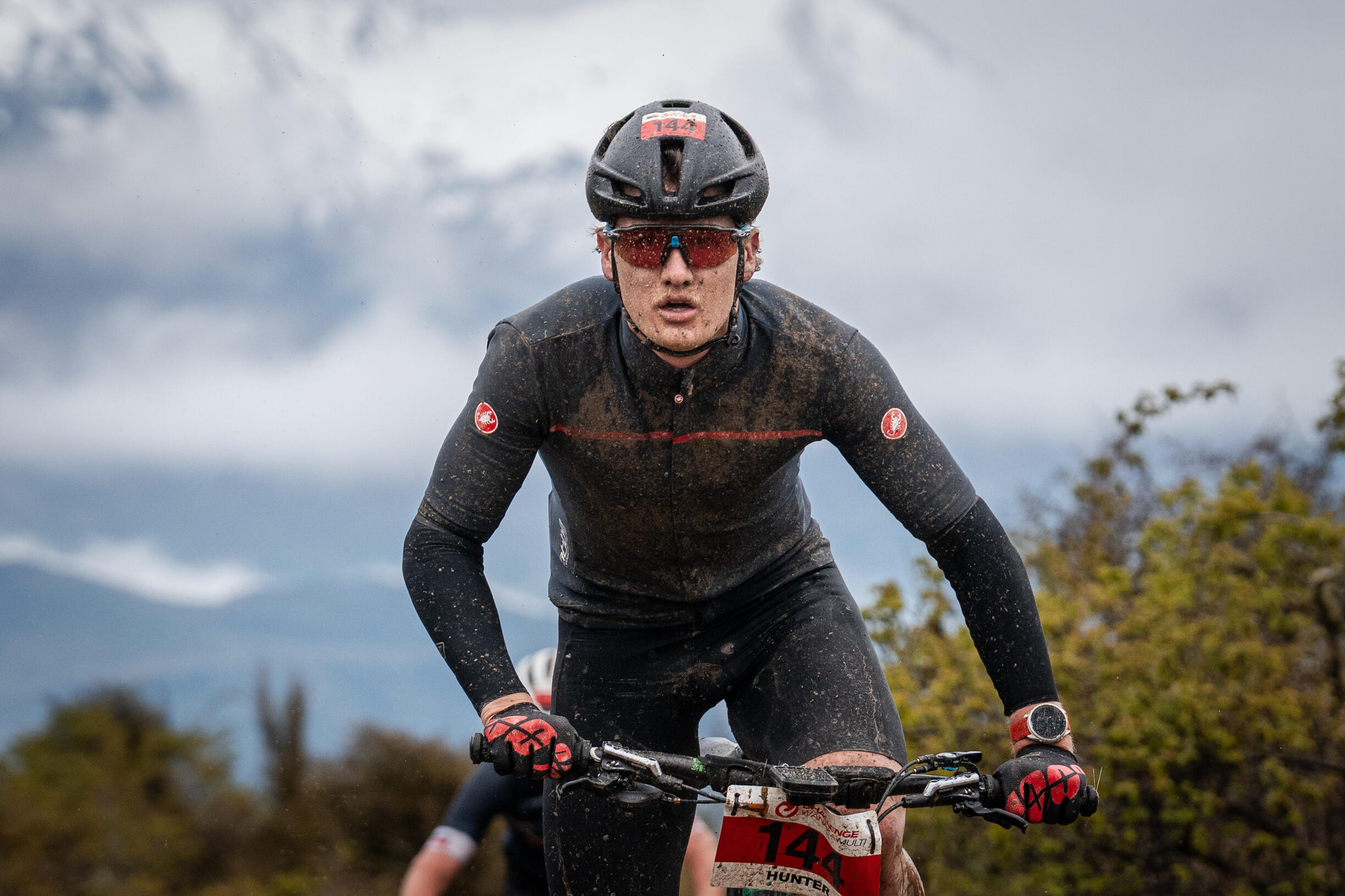 Mountain biker covered in mud racing downhill on technical trail, wearing black kit and full-face helmet, intense focused expression
