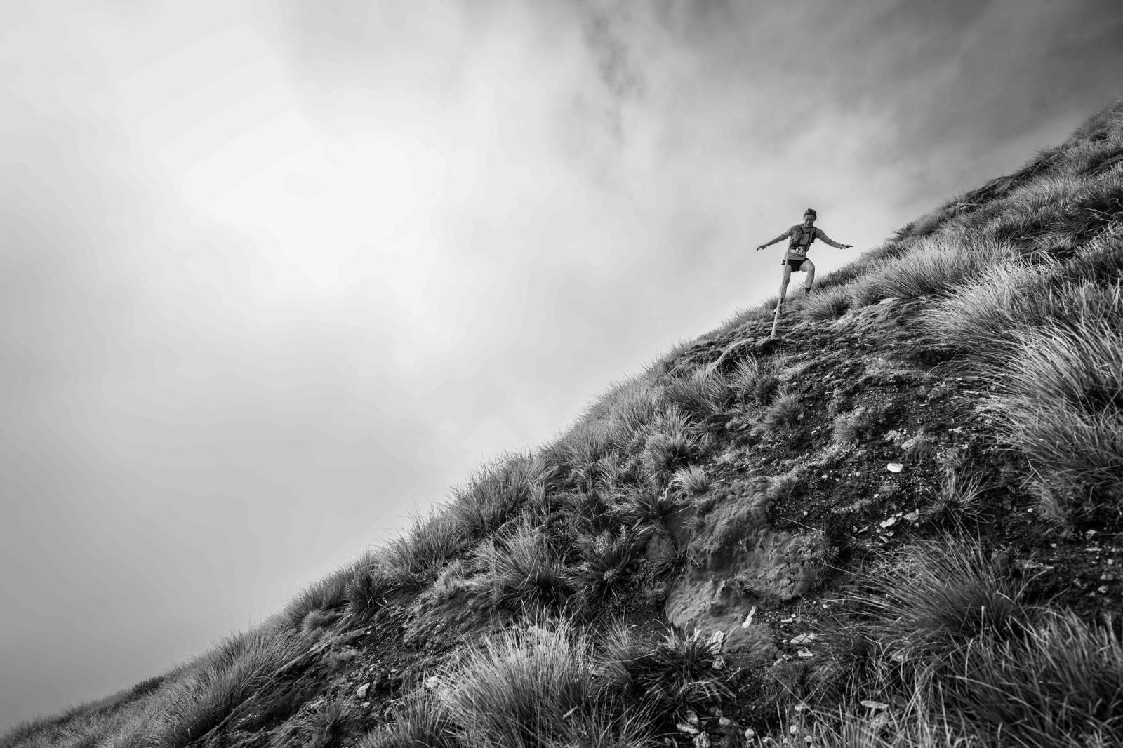 Runner Treble Cone Wanaka New Zealand Silhouette of trail runner descending steep grassy mountain slope in dramatic black and white, stormy clouds overhead creating moody atmosphere