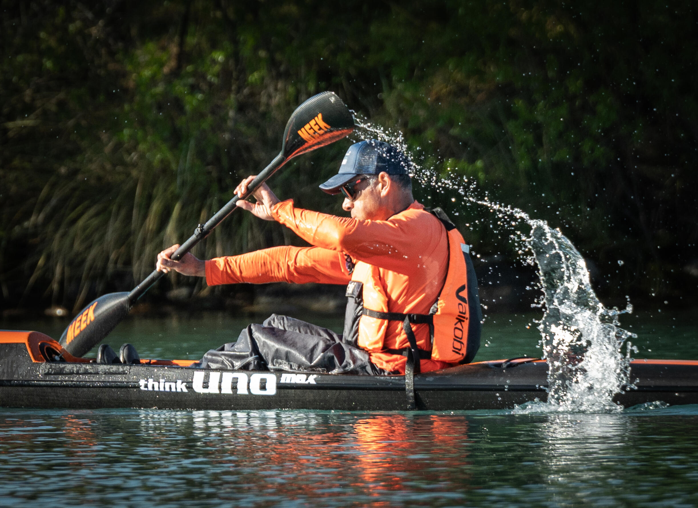 Nathan Faavae Meridian Twizel Hard Labour Kayaker in orange life vests paddling tandem canoe, water splashing from paddle stroke, lush green forest background