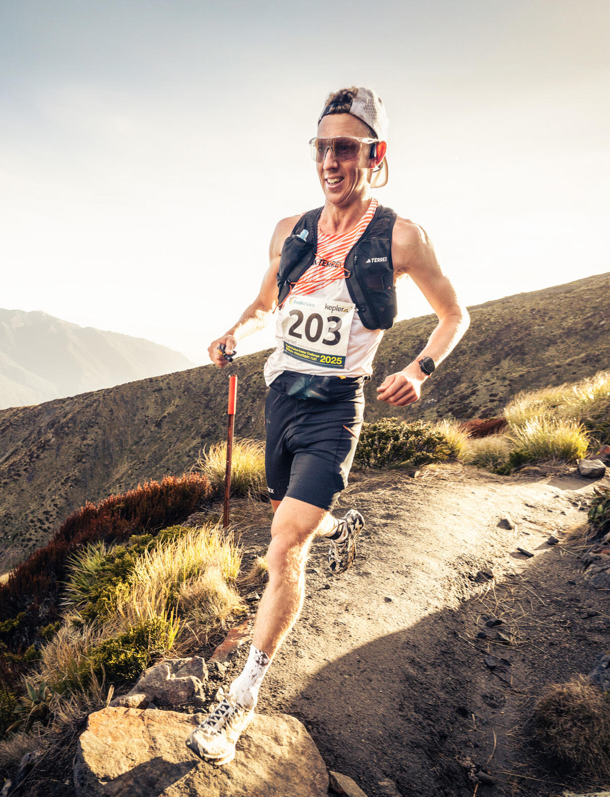Daniel Jones Kepler Challenge New Zealand Trail runner with race number 203 ascending a mountain trail at golden hour, wearing sunglasses and hydration vest against dramatic hillside backdrop