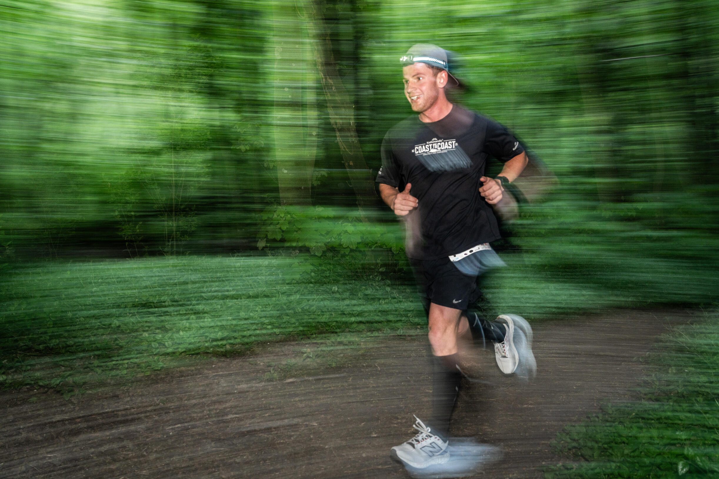 Arrowtown Backyard Ultra Trail runner in black gear and cap captured with motion blur running through lush green forest, dynamic panning photography technique