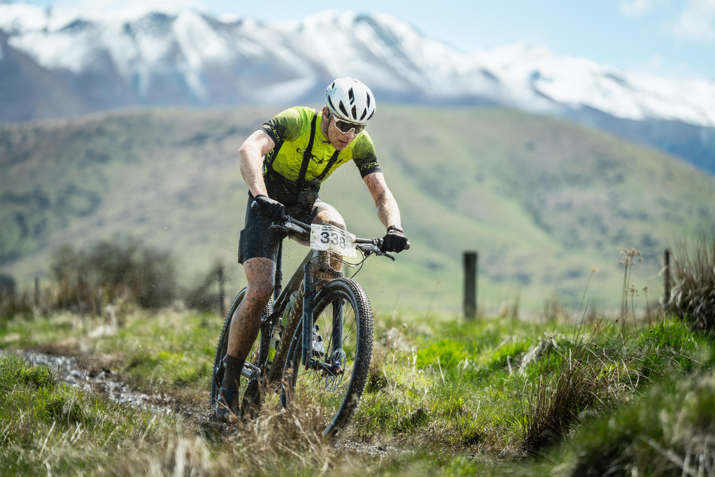 Meridian Twizel Hard Labour Mountain biker in bright yellow jersey racing through muddy trail with race number 383, snow-capped peaks and alpine meadow in background